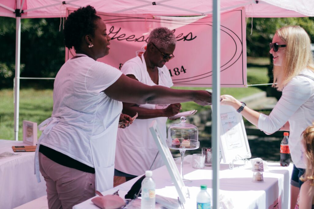 Women wearing serving a customer in outdoor setting. Pink marketing signage is in the background.