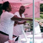 Women wearing serving a customer in outdoor setting. Pink marketing signage is in the background.